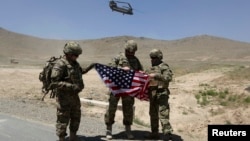 Tentara NATO berpose sambil memegang bendera Amerika sementara helikopter Chinook lepas landar seusai serah terima pengalihan tanggung jawab keamanan di akademi militer di luar Kabul (18/6). Afghanistan menangguhkan perundingan bilateral dengan AS, menyusul rencana dialog AS-Taliban.