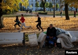 FILE - Youths train behind a homeless man sitting on a bench at the Alexander Garden on a sunny autumn day in St. Petersburg, Russia, Oct. 16, 2018.