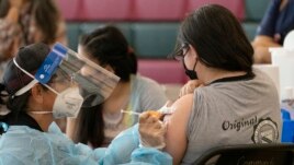 Sisters Guadalupe Flores, 15, right, and Estela Flores, 13, left, from East Los Angeles, get vaccinated with the Pfizer's COVID-19 vaccine by licensed vocational nurse Rita Orozco, far left, at the Esteban E. Torres High School in Los Angeles, Thursday, M