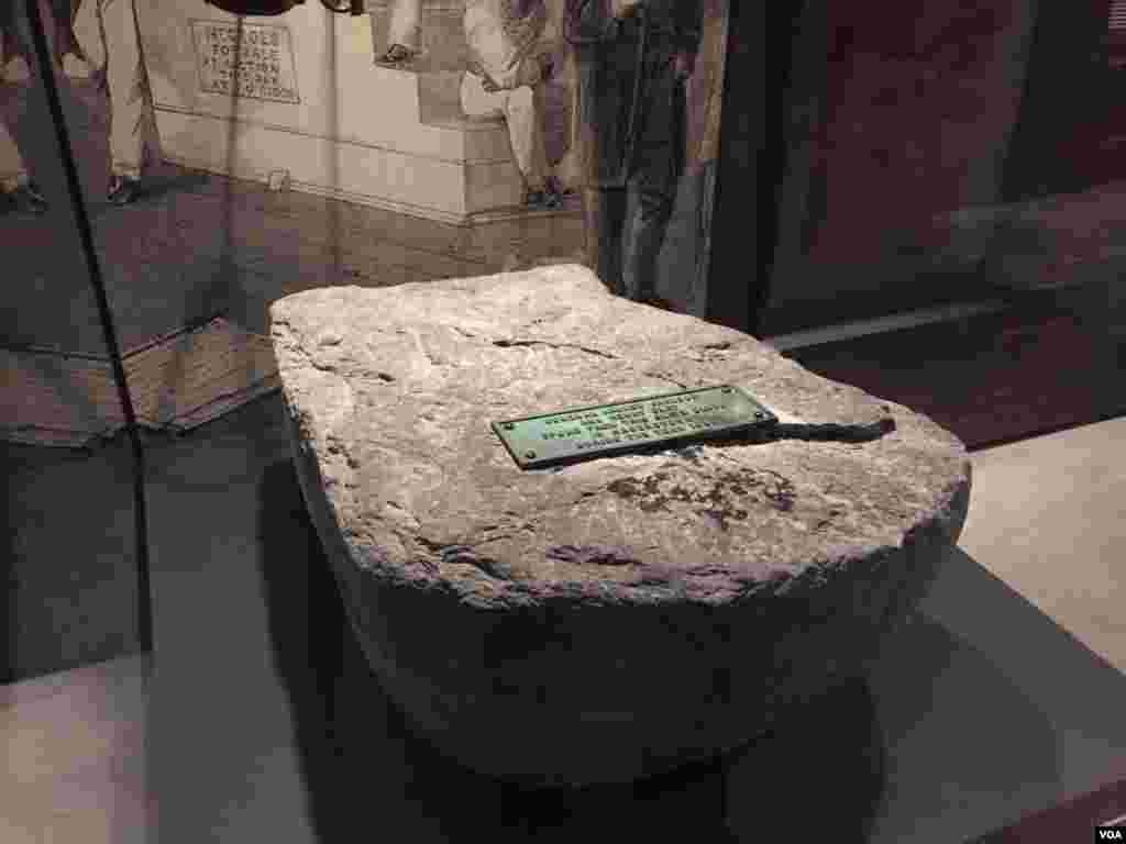 A fragment of a stone slave auction block from Hagerstown, Maryland, is displayed at the Smithsonian National Museum of African American History & Culture in Washington, D.C., with a scene recreating a slave auction seen in the background. (C. Simkins/VOA