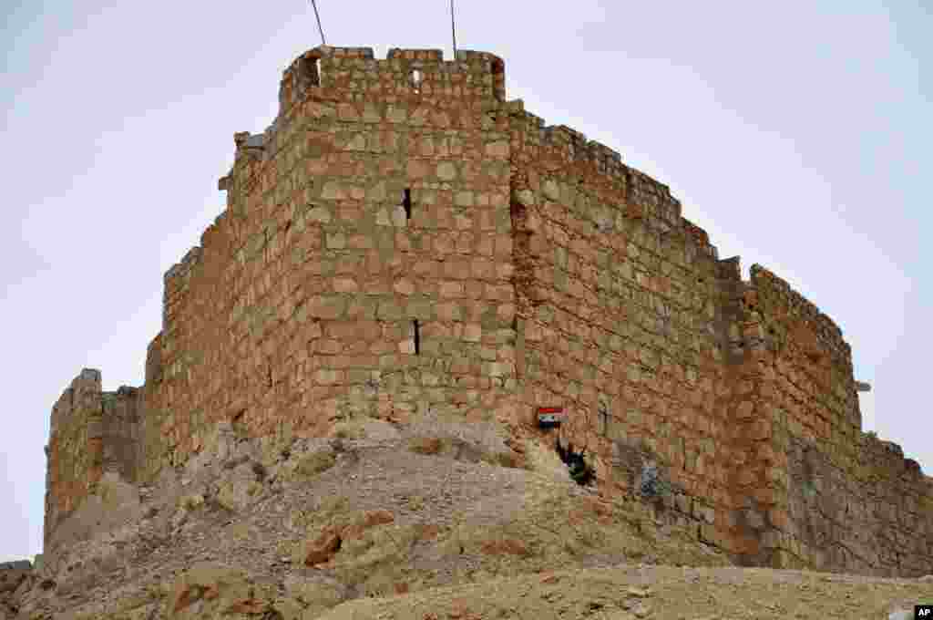 In this photo released by the Syrian official news agency SANA, a Syrian soldier holds a Syrian national flag in front of the Palmyra citadel, March 27, 2016.