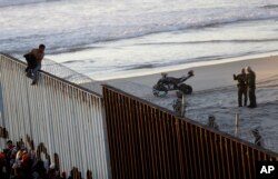 지난달 중남미 출신 이민자 행렬 캐러번이 미국과 멕시코 국경에 도착한 가운데, 한 이민자가 국경담장에 올라타자 미국 국경수비대가 그 모습을 지켜보고 있다.