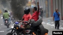 2 neg kap pase sou yon motosiklèt nan yon ri nan Pòtoprens kote kawotchou ap boule. Pòtoprens, Ayiti, 10 jiyè 2019. Foto: REUTERS/Andres Martinez Casares