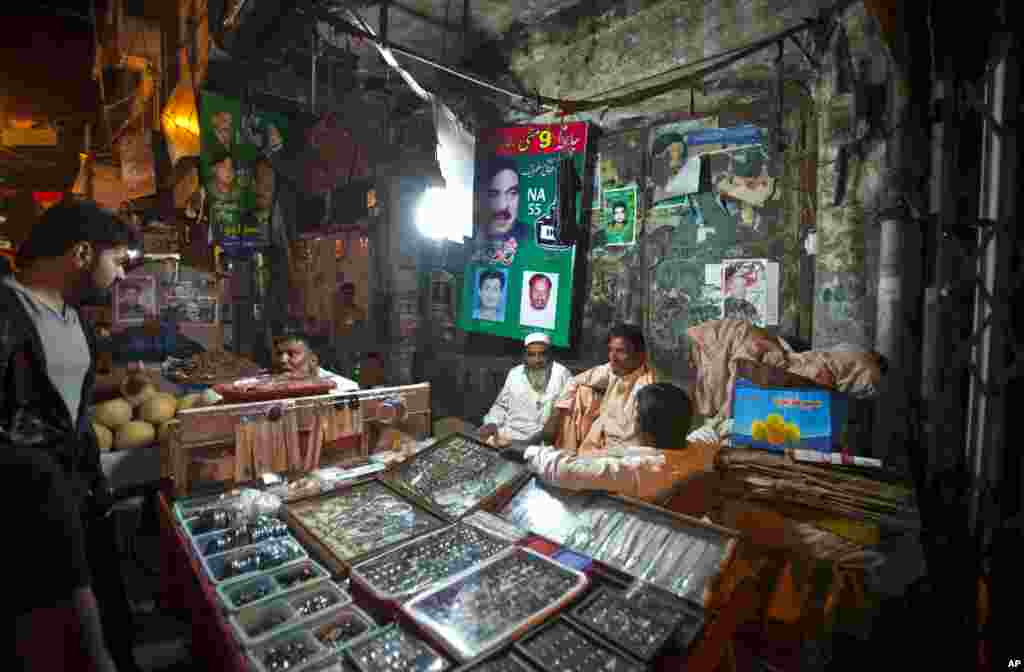 A vendor talks to a customer as others chat at a stall next to posters of various candidates for the upcoming election pasted on a wall in Rawalpindi, Pakistan, May 8, 2013.