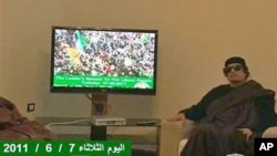 In this image from a TV broadcast by Libyan television, Libyan leader Moammar Gadhafi sits next to a TV monitor showing a strapline at the bottom in English, reading 'The Leader's speech to the Libyan people 07 06 2011