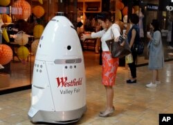 FILE - Sophie Li of Cupertino, California, offers to dance with a K5 robot made by Knightscope Inc., at Westfield Valley Fair shopping center in San Jose, California, June 26, 2017. In Silicon Valley, people can eat a pizza made largely by a robot, have hotel toiletries delivered by a robot, drink a frothy cappuccino made by a robot and shop at a mall with robot security.