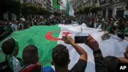 FILE - Protestors carry a large flag and chant slogans during a demonstration against the country's leadership, in Algiers, April 12, 2019.