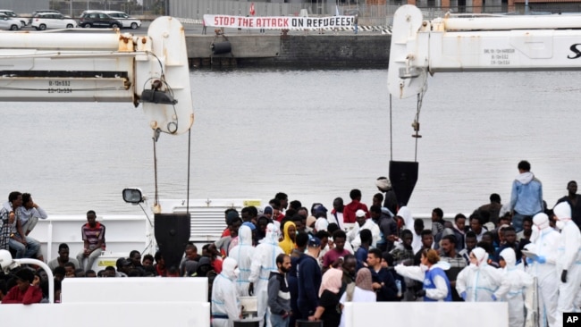 Activistas levantan una pancarta que dice "Detengan el ataque a los refugiados" mientras inmigrantes aguardan para desembarcar del barco "Diciotti" de la Guardia Costera italiana, en el puerto siciliano de Catania, en el sur de Italia, el miércoles 13 de junio de 2018.