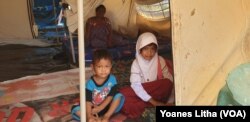 Saskiah (7) seusai pulang sekolah duduk diatas kasur di dalam salah tenda ditemani adik dan ibunya, 22 Juli 2019. (Foto: Yoanes Litha/VOA)