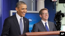 FILE - President Barack Obama, left, shares a laugh with White House press secretary Jay Carney, right, as the president makes a surprise visit to the Brady Press Briefing Room in Washington.