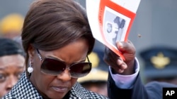 FILE - A bee hovers near Zimbabwe's first lady Grace Mugabe's head, as an aide comes to her assistance, during a funeral service in Harare, May, 13, 2017. Mugabe faces charges of assault after an incident at a plush Johannesburg, South Africa, hotel.