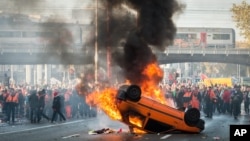 Người biểu tình đứng đằng sau một chiếc xe đang cháy trong một cuộc biểu tình của công đoàn quốc gia ở Brussels, 6/11/2014.