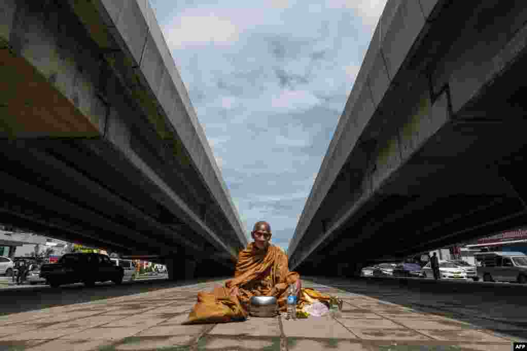 ព្រះសង្ឃពុទ្ធសាសនាមួយអង្គកំពុងអង្គុយនៅលើដី នៅចន្លោះអាគារពីរ នៅពេលព្រះអង្គរង់ចាំចង្ហាន់នៅក្នុងទីក្រុងបាងកក។