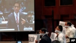 Lawmakers from the opposition wearing gags and holding protest posters against the new Communications Act as the screen shows a live image of lawmaker Mauro Andino presenting his proposal.