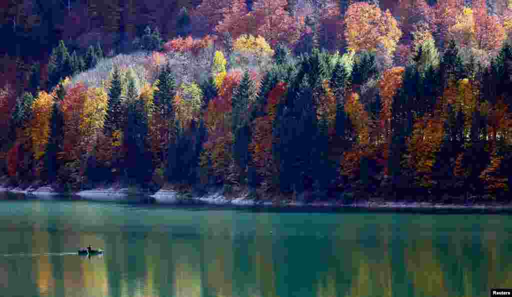 Autumnal trees are reflected in the water as a fisherman rows his boat during a sunny day at the Sylvenstein barrier lake in Germany.