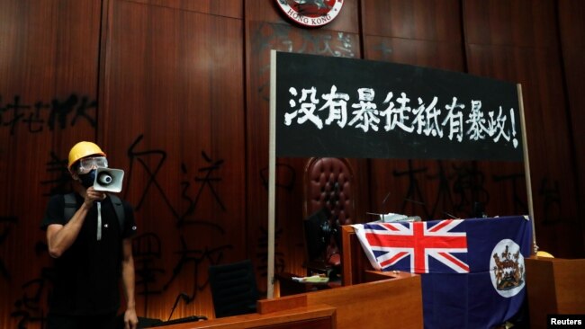 一名进入香港立法会的抗议者站在挂在立法会内的港英旗帜旁使用扩音喇叭讲话。（2019年7月1日）
