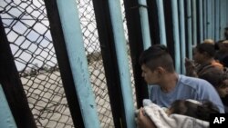 Un miembro de la caravana proveniente de Centroamérica, cargando un bebé, mira a través de una reja de alambre como un grupo se reúne a la frontera, mientras él espera en una playa en Tijuana, México. 29 de abril, 2018.