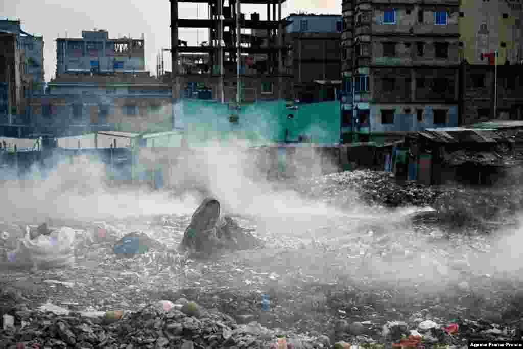 A woman searches reusable items from a pile of trash as smoke rises from a nearby area in Dhaka, Bangladesh.