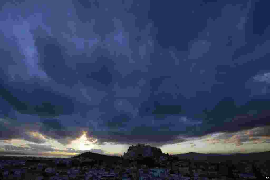 A sunset seen through heavy cloud cover behind the ancient Acropolis hill in Athens, Greece.