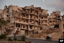 In this Sept. 20, 2018 photo, motor cycles ride past buildings destroyed during the fighting in the northern town of Ariha, in Idlib province, Syria.