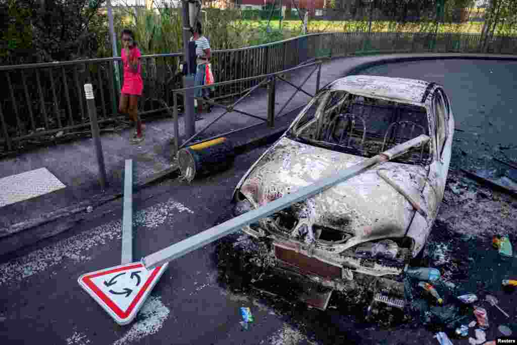 A girl walks past a street barricade with a burned car after unrest triggered by COVID-19 curbs in Fort-De-France, Martinique.