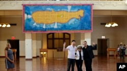 Bono, right, and The Edge, left, of the Irish rock band U2, flank Yoko Ono, the widow of John Lennon, at the unveiling of a giant tapestry depicting the island of Manhattan as a yellow submarine piloted by a waving Lennon at Ellis Island, July 29, 2015, 