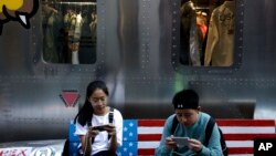 FILE - Shoppers sit on a bench with a decorated with U.S. flag browsing their smartphones outside a fashion boutique selling U.S. brand clothing at the capital city's popular shopping mall in Beijing.