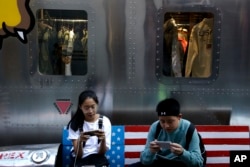 FILE - Shoppers sit on a bench with a decorated with U.S. flag browsing their smartphones outside a fashion boutique selling U.S. brand clothing at the capital city's popular shopping mall in Beijing, Sept. 24, 2018.