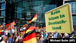 Pendukung partai AfD Jerman mengibarkan bendera di depan stasiun kereta di Berlin, Jerman, Minggu, 27 Mei 2018. Partai AfD yang memanfaatkan sentimen anti-migran untuk mendapatkan kursi di parlemen, melakukan protes pada bulan Mei menentang pemerintah sambil membawa poster bertuliskan "tidak boleh ada Islam di Jerman". 