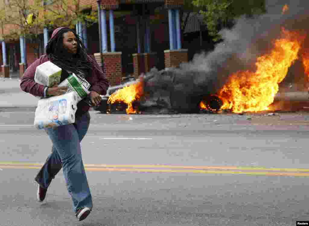 A woman with goods looted from a store runs past burning vehicles during clashes in Baltimore, Maryland, April 27, 2015. 