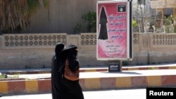 IŞİD'in kurduğu sözde halifelik devletinin merkezi Rakka'da kadınların tamamen çarşaf giymesi yönünde Kuran'dan alıntı veren afişin altında yürüyen iki kadın.