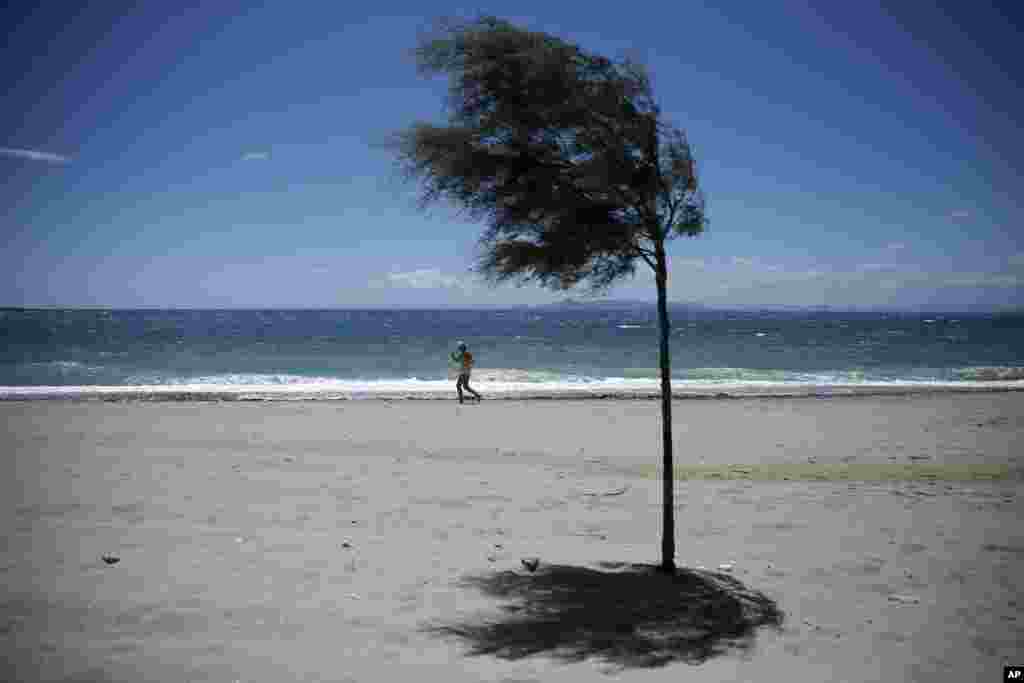 A woman jogs at the seaside of Alimos in front of a tree during a windy day, Athens, Greece.