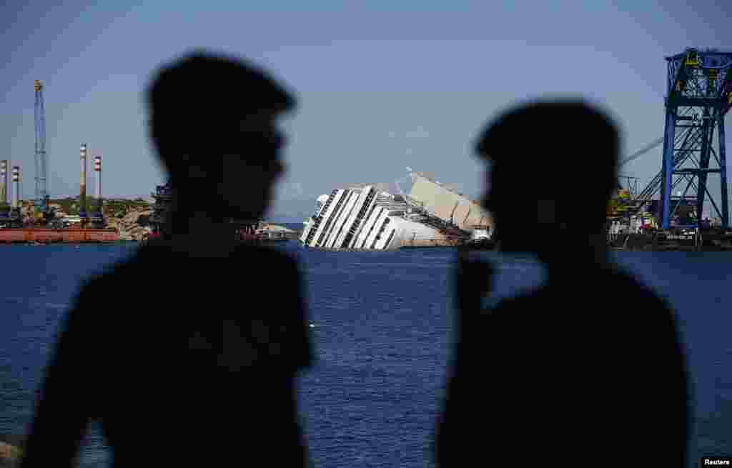 Siluet dua orang pria dengan latar belakang kapal pesiar Costa Concordia yang terbalik di pelabuhan Giglio, Italia.