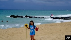 Seorang gadis berjalan di pantai berpasir memegang bunga matahari dan sandal jepitnya di distrik Condado San Juan, Puerto Rico, Rabu, 28 Agustus 2019. (Foto: AP)