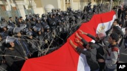Para pengunjuk rasa Mesir memasang spanduk raksasa berwarna bendera kebangsaan Mesir di barikade kawat berduri yang dipasang di depan tentara anti huru-hara yang berjaga di depan pintu masuk istana Kepresidenan Mesir di Kairo, 25 Januari 2013. (AP Photo/Amr Nabil)