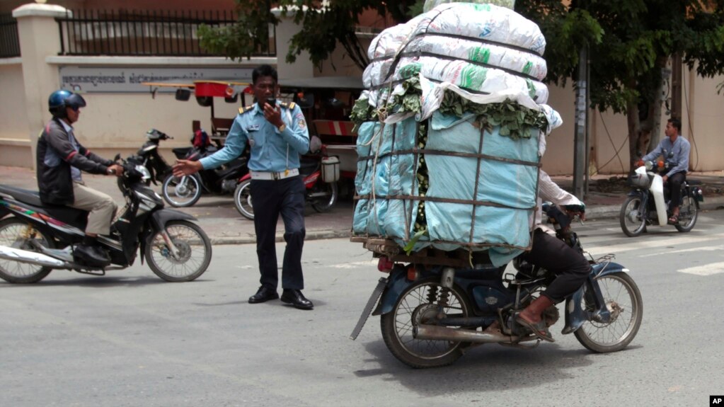 A Cambodian man, right, drive his motorbike overloaded with vegetable sacks as he passes a traffic police officer, center, in Phnom Penh, Cambodia, Tuesday, Aug. 25, 2015. (AP Photo/Heng Sinith)