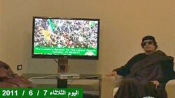In this image from a TV broadcast by Libyan television, Libyan leader Moammar Gadhafi sits next to a TV monitor showing a strapline at the bottom in English, reading 'The Leader's speech to the Libyan people 07 06 2011,' during a meeting with unidentified