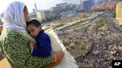 An Egyptian mother hugs her child as she watches thousands of Egyptian protesters gather at Tahrir square in Cairo, Egypt, Sunday, Jan. 30, 2011, with the ruling National Democratic party building burned at top right behind the red colored Egypt museum.