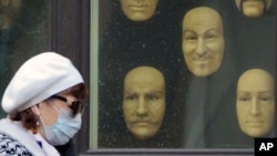 A woman wearing a face mask to protect against coronavirus walks past wax faces displayed in a window of a wax museum in St.Petersburg, Russia, Monday, May 4, 2020.