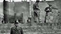 图为1961年10月7日一名西柏林士兵站立在分割东、西柏林的混凝土墙边。他身后为东柏林工人正在加高围墙