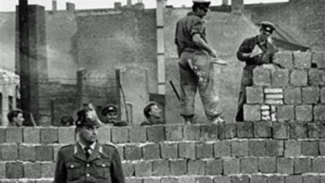 图为1961年10月7日一名西柏林士兵站立在分割东、西柏林的混凝土墙边。他身后为东柏林工人正在加高围墙