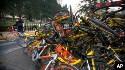 FILE - A woman rides a bicycle past Mobike and Ofo branded shared bicycles piled on a traffic median in Beijing, Nov. 21, 2017.