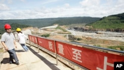 FILE PHOTO- A Chinese engineer, left, walks by a fence with Chinese slogans reading: Safety first, highest quality and quantity near an entrance of a dam construction site by China National Heavy Machinery Corporation on the Tatay River in Koh Kong province. 