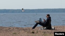 Seorang pria bekerja dengan menggunakan smartphone dan laptopnya, sambil duduk di pantai Teluk Finlandia di tengah pandemi Covid-19 di Saint Petersburg, Rusia, 27 Mei 2020. (REUTERS / Anton Vaganov)