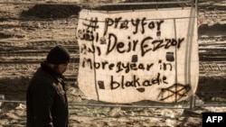FILE - A man walks past a banner reading "Pray for Deir ez-Zor" at a migrant camp in Calais, France, Dec. 7, 2015. Years of conflict between Syrian government forces and Syrian opposition groups, and the emergence of extremist groups like Islamic State, have forced hundreds of thousands of people to flee from Deir ez-Zor province. 