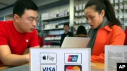 An employee works on a laptop computer as he talks with a customer near a sheet showing accepted methods of payment, including, beginning Thursday, Apple Pay, top left, at an Apple Store in Beijing, Feb. 18, 2016.