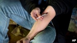 FILE - A man injects heroin into this arm under a bridge along the Wishkah River at Kurt Cobain Memorial Park in Aberdeen, Washington, June 13, 2017.