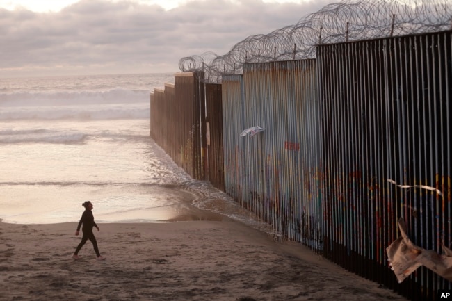 FILE - A woman walks on the beach, Jan. 9, 2019, next to the border wall topped with razor wire in Tijuana, Mexico.