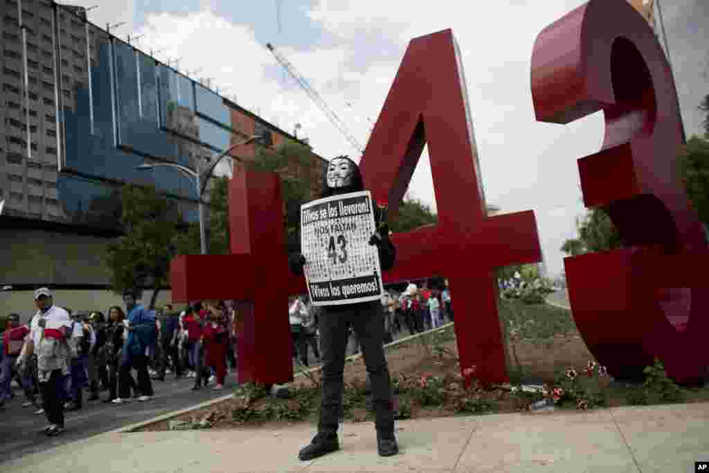 Demonstran bertopeng berpose dengan papan bertuliskan &quot;Kami kehilangan 43 orang. Mereka menangkapnya hidup-hidup. Kami ingin mereka kembali hidup-hidup&quot; di depan monumen yang baru didirikan untuk para mahasiswa keguruan yang hilang dari negara bagian Guerrero, dalam pawai Hari Buruh di Mexico City (1/5). ​(AP/Rebecca Blackwell)