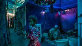 A young Rohingya refugee girl walks past an elderly man sitting inside a tent at a refugee camp alongside the banks of the Yamuna River in the south-eastern borders of New Delhi. (AP Photo/Altaf Qadri)